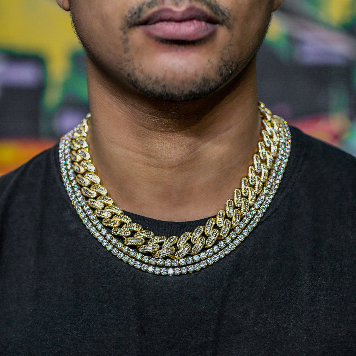 Close-up of man's neck/chest with layered chains: 12mm iced cuban + thinner gold cuban + diamond tennis. Black tee, graffiti wall.